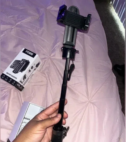 Hand holding a black extendable selfie stick with phone mount, box labeled P15 MINI on bed